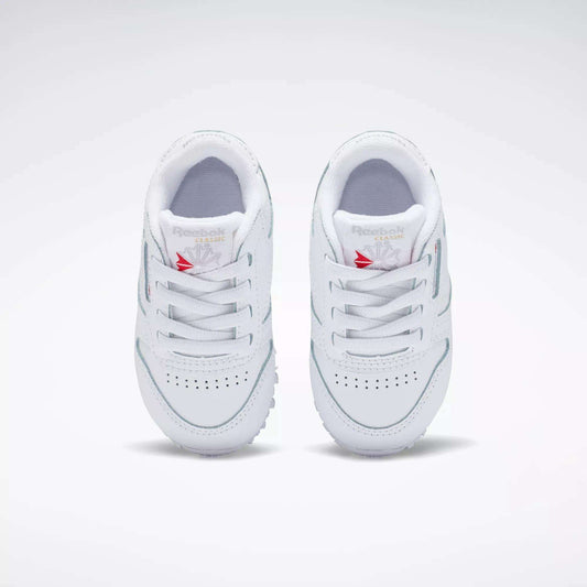 A pair of Kids' Classic Leather Shoes - Baby & Toddler by Reebok in white, featuring laces, toe perforations, and a red Reebok logo on the tongue, shown from above on a plain light background—ideal for toddlers’ everyday style.