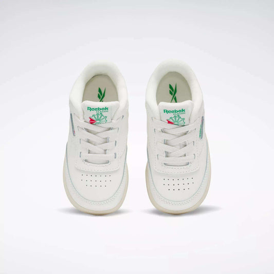 A pair of Reebok Kids' Club C Shoes for toddlers in white with green and pink accents, inspired by '80s style, shown from above on a plain background. Both shoes feature the Reebok logo and a leaf graphic on the tongue.