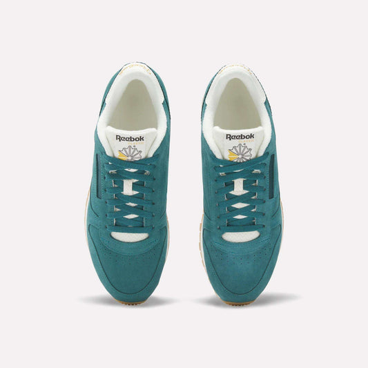 A pair of Classic Leather Shoes by Reebok in teal suede with white accents, shown from above. The image highlights the laces, gold and white branded tongue, beige outsoles, and a light background.