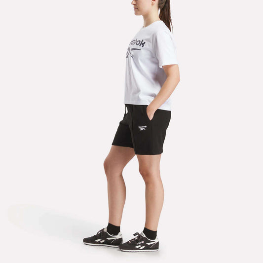 A woman stands sideways against a plain white background wearing a white Reebok t-shirt, black Women’s Pintuck Long Shorts by Reebok with the logo, black socks, and black sneakers.