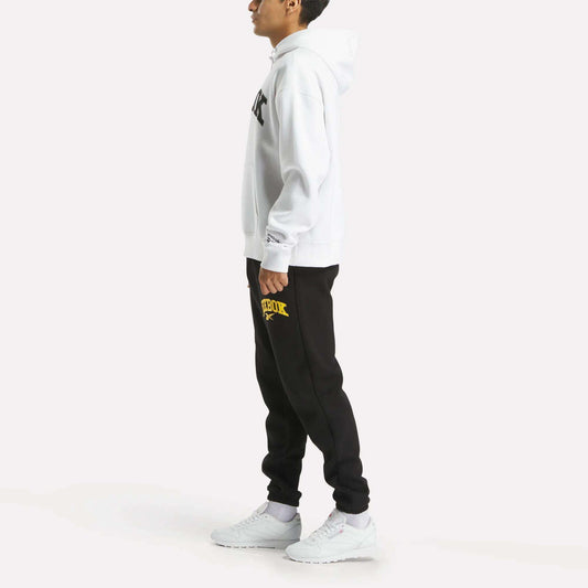 A person in profile wears a white Reebok Men's Hoodie, black sweatpants with a yellow Reebok logo, and white sneakers, standing against a plain white background.