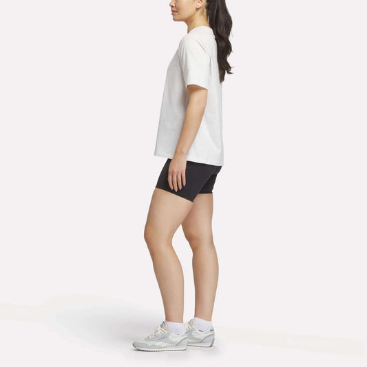A woman stands in profile wearing the Reebok Women's Lawn Club T-Shirt, black shorts, white socks, and gray sneakers against a plain white background.