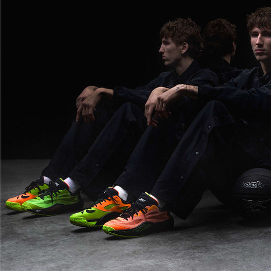 A person in dark clothing sits on the floor, arms on knees, wearing mismatched neon green and orange Reebok Engine A 26 Basketball Shoes. Their reflection and a black Reebok basketball are seen in the background, showcasing style and stability.