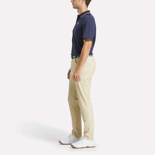 A man in profile wears the Reebok Men's BDC Signature Polo Shirt, beige pants, a white belt, and white shoes, standing against a plain white background.