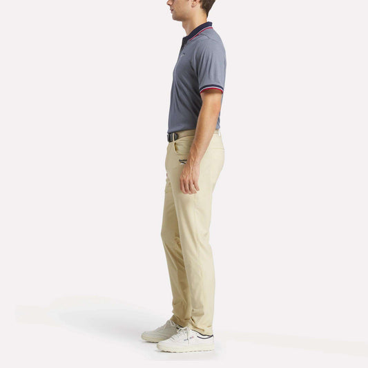 A man stands in profile wearing a men's Reebok Country Club Oxford Pique Polo Shirt, beige pants, white sneakers, and a black belt against a plain white background.