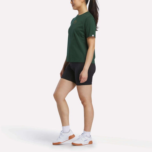 A woman with a ponytail stands sideways on a white background, wearing the Reebok Women's Fitted Graphic T-Shirt in green, paired with black shorts, white socks, and white sneakers with gum soles.