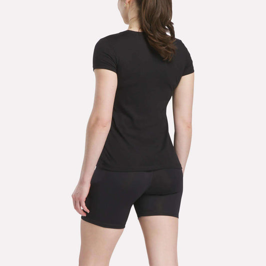 A person with long brown hair in a ponytail is standing and facing away, wearing a Reebok Women's T-Shirt and black shorts against a plain white background.
