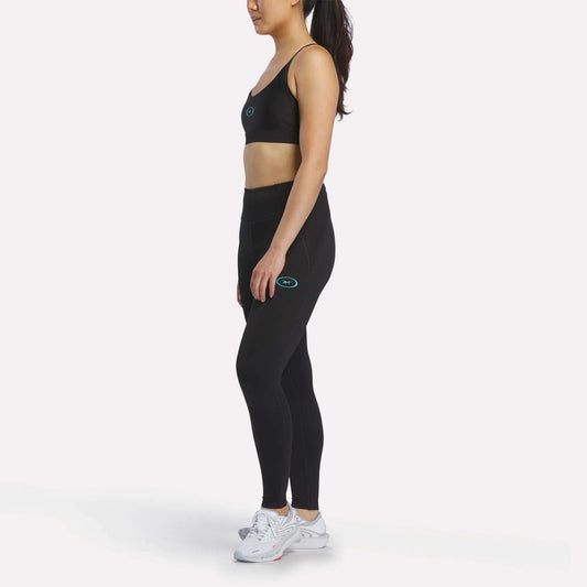 A woman in a black Reebok sports bra and Women's Speed HR High-Rise Leggings stands sideways on a white background, hair in a ponytail. Both clothing pieces feature a small blue and white Reebok logo. She pairs the outfit with white sneakers.