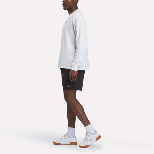 A man stands in profile against a plain white background wearing a Reebok Men's Long Sleeve T-Shirt in white, black shorts, white socks, and white Reebok sneakers with gum soles.