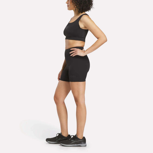 A woman stands in profile against a plain white background, wearing a black Women's Reebok x F45 Training Sports Bra, paired with black high-waisted biker shorts and black running shoes.