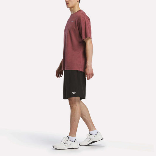 A person stands in profile against a plain white background, wearing a maroon Reebok Men's Director Short Sleeve Top, black shorts, white socks, and white athletic shoes.