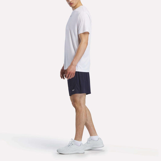 A man stands sideways wearing a plain white t-shirt, Reebok Men's Inclination Shorts, and white sneakers against a plain light background.