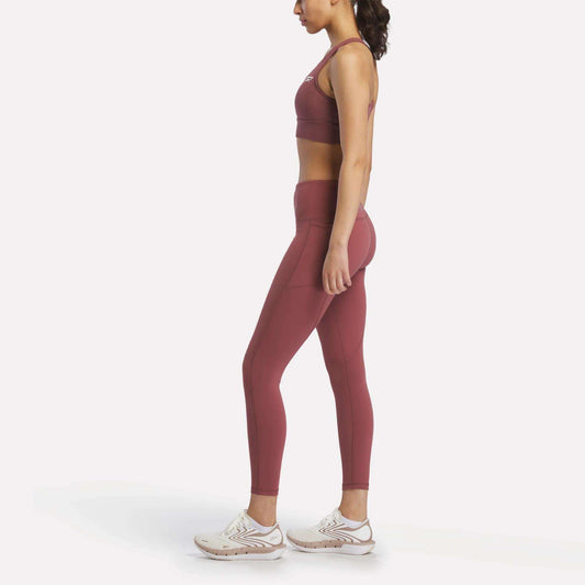 A woman in profile faces left, wearing Reebok Women's Odyssey High-Rise 7/8 Leggings and a matching sports bra, paired with white athletic shoes, set against a plain white background.