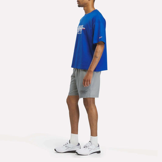 A person stands sideways wearing a blue Reebok x F45 Training Graphic T-Shirt, gray shorts, white crew socks, and white sneakers against a plain white background.