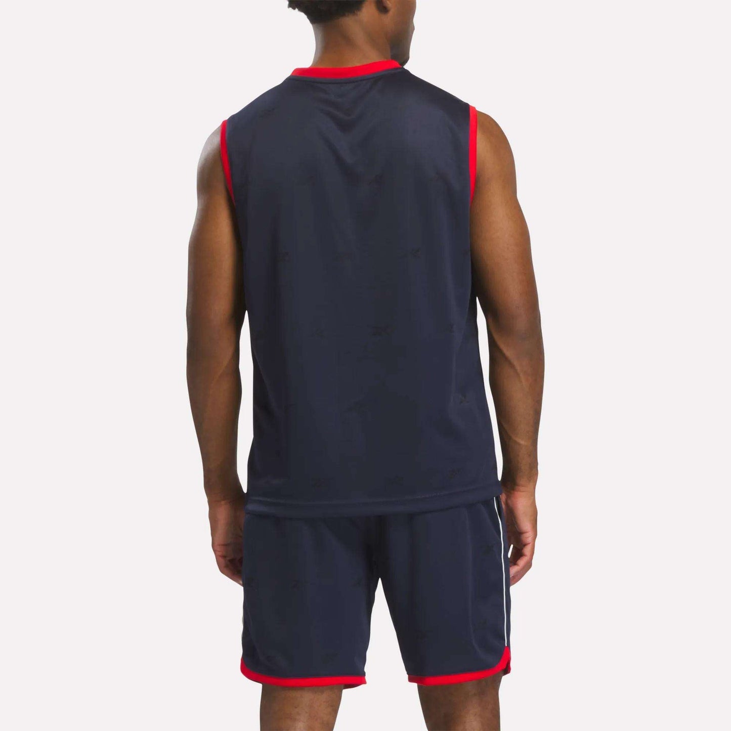 A man stands with his back to the camera, wearing a Reebok Men's United Jersey Tank and matching shorts in navy blue with red trim, resembling a soccer sports jersey.