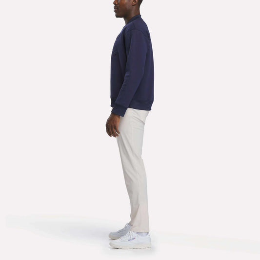 A person stands sideways against a plain white background, wearing the Reebok Men's Signature Golf Crewneck Sweatshirt in navy, paired with light-colored pants and white sneakers.