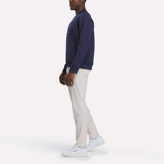 A person stands sideways against a plain white background wearing a navy sweatshirt, white sneakers, and Reebok Men's Favorite 5-Pocket Relaxed Fit Golf Pants in a wrinkle-resistant fabric.