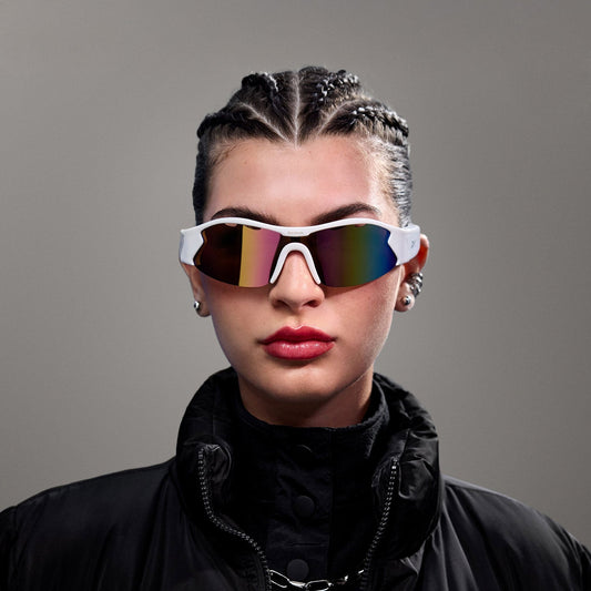 A person with braided hair wearing Reebok Flash Smart Audio Sunglasses with polarized lenses, a black puffer jacket, and bold red lipstick poses against a plain gray background.