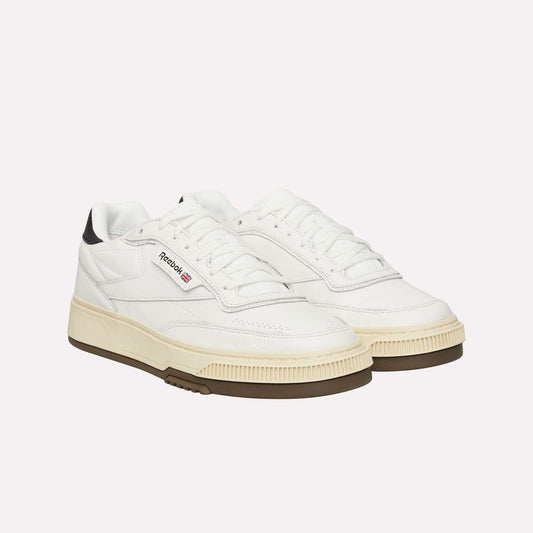 A pair of white Reebok sneakers with black detailing on the heel, off-white midsoles, gum rubber outsoles, and the Reebok logo on the side. The shoes have a retro design with lace-up fronts.