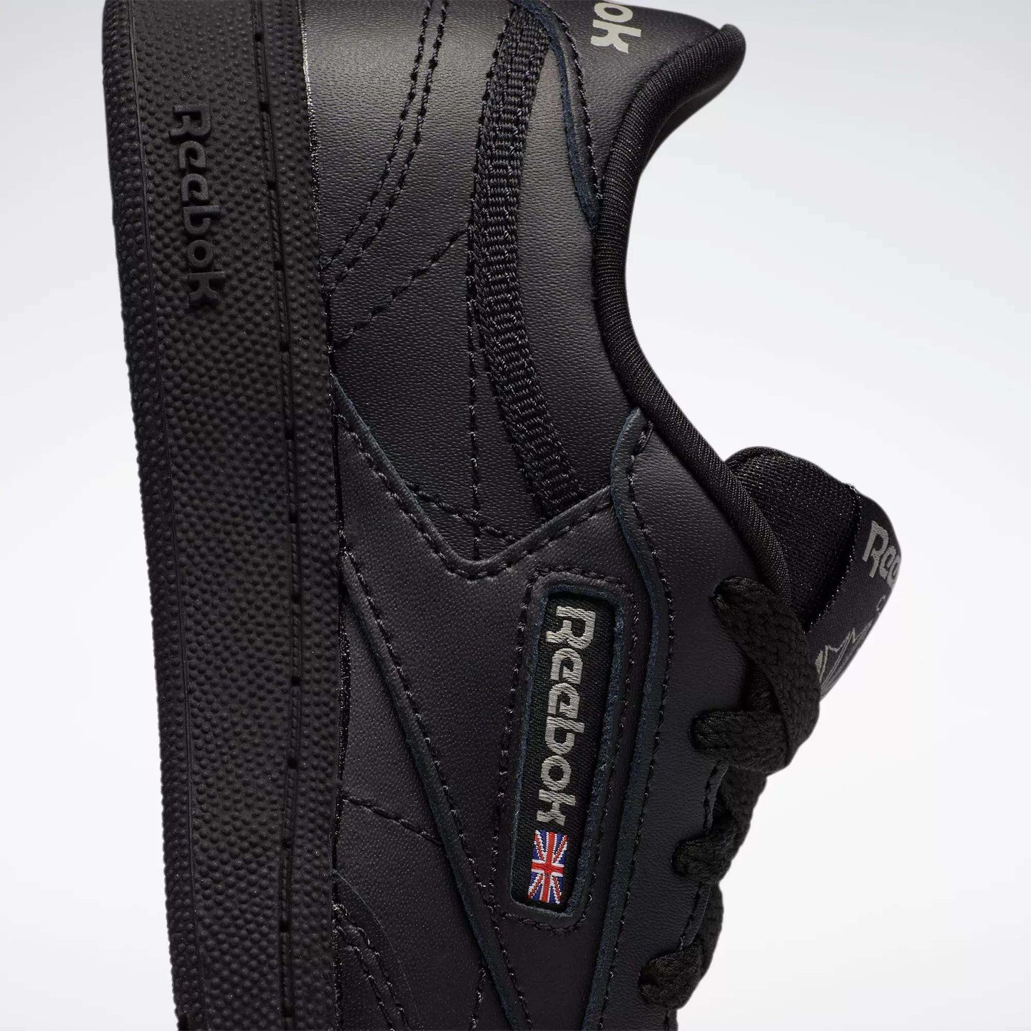 Close-up of the Reebok Kids' Club C Shoes for little kids, featuring a black leather upper with the Reebok logo and a small British flag patch on the side. The shoe is slightly tilted to show its side profile and textured sole.