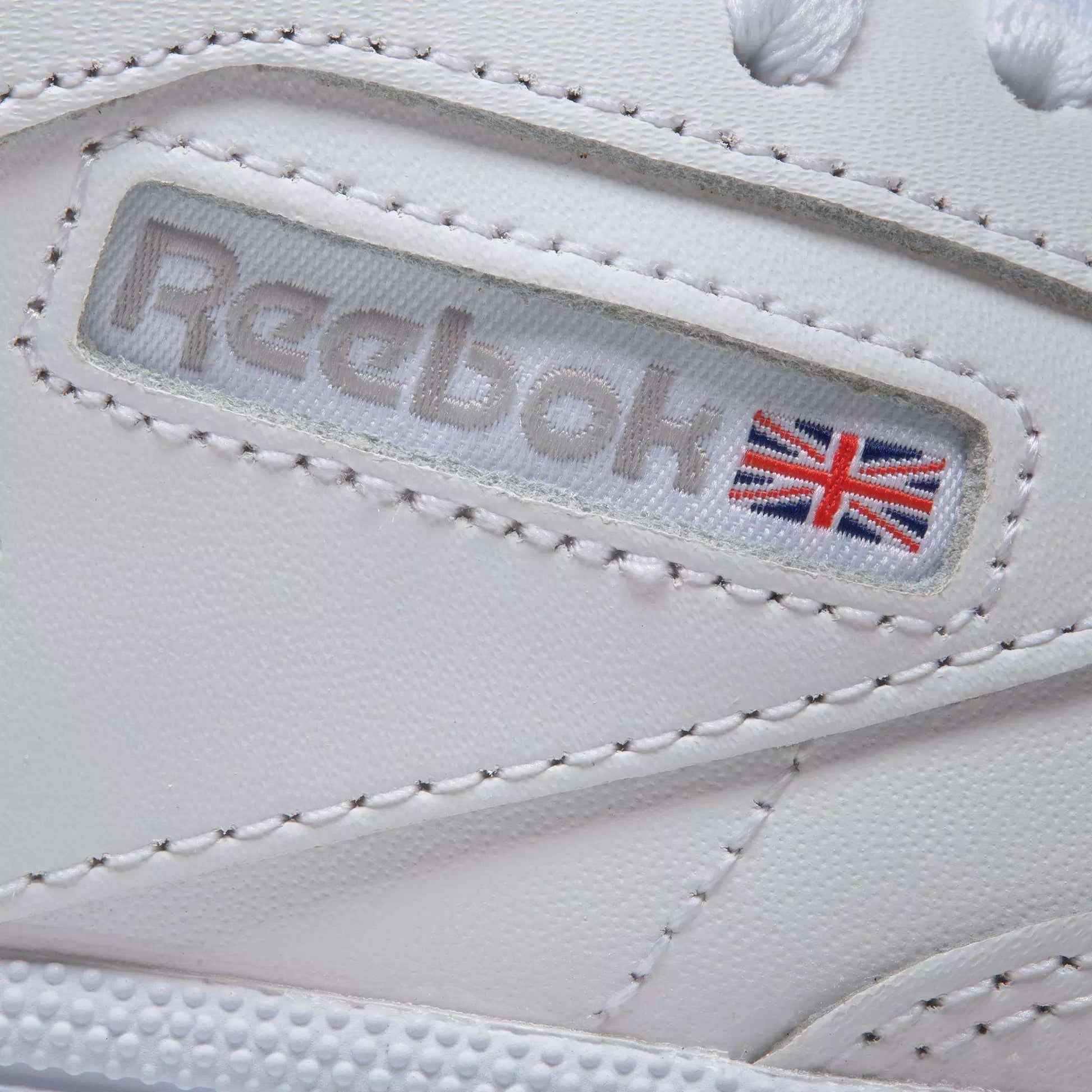 Close-up of Reebok Women's Club C 85 Shoes in white, featuring a stitched Reebok logo and Union Jack flag on a leather panel with visible stitching—showcasing iconic style and detailed craftsmanship.