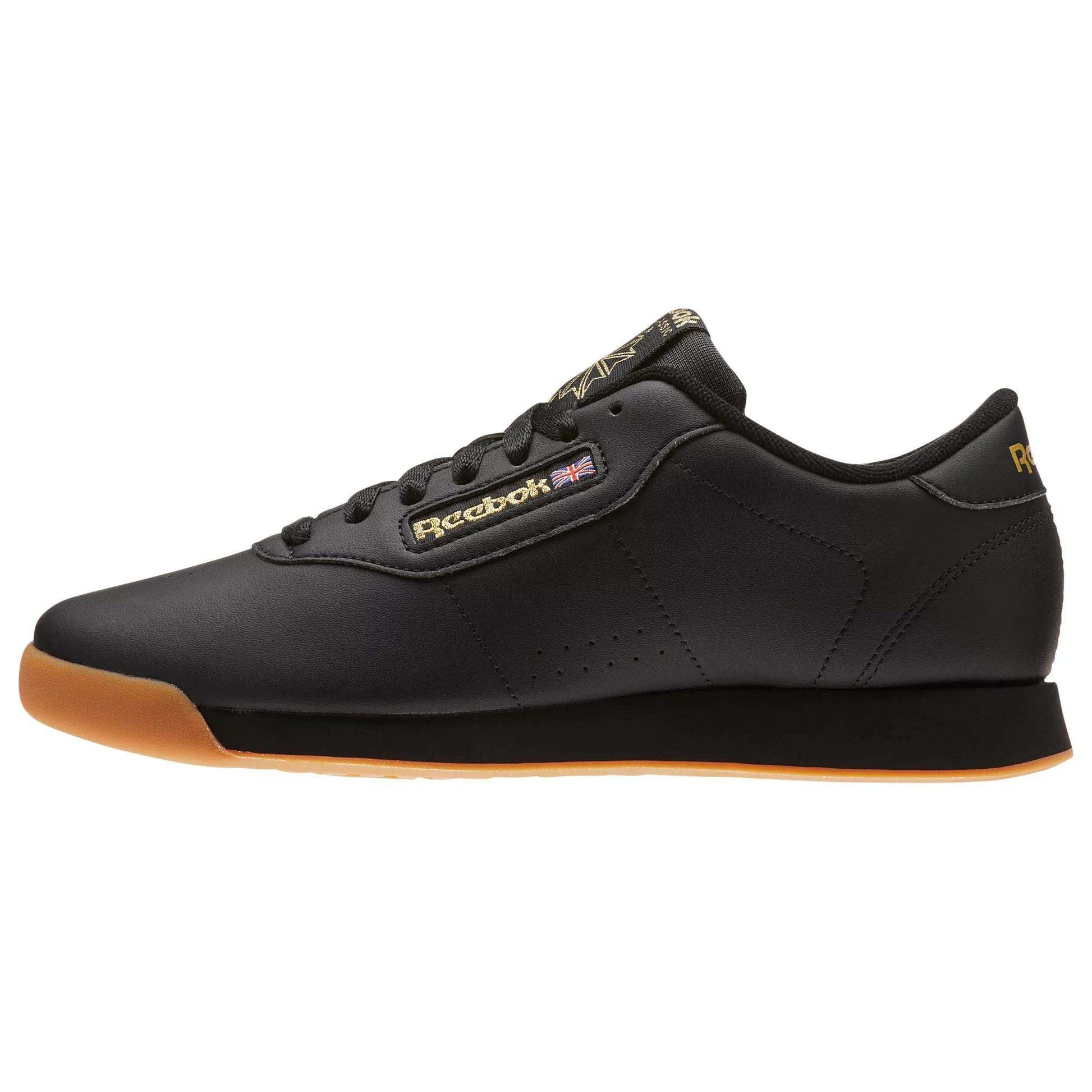 The Reebok Women's Princess Shoes are black sneakers with a gum sole, gold branding, and a small Union Jack flag. They feature black laces, a low-cut design, and provide cushioned comfort in a heritage style.