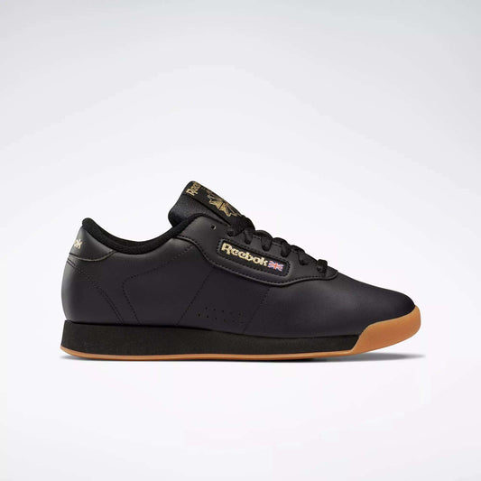 The Reebok Women's Princess Shoes in black feature a gum sole, black laces, and gold Reebok branding on the side, tongue, and heel. Combining cushioned comfort with heritage style, they're displayed against a plain light gray background.