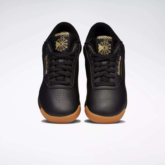 A pair of Reebok Women's Princess Shoes in black with gum soles and gold logos, featuring a cushioned design, are placed side by side on a plain light background, facing forward.