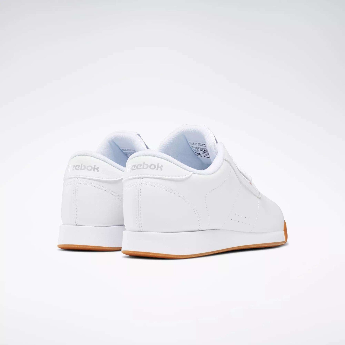 The Reebok Women's Princess Shoes, shown from back and side angles on a white background, feature a classic low-top design, gum-colored soles, and subtle cushioning for everyday comfort.