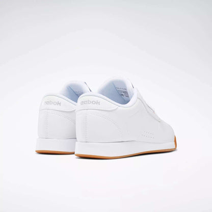The Reebok Women's Princess Shoes, shown from back and side angles on a white background, feature a classic low-top design, gum-colored soles, and subtle cushioning for everyday comfort.