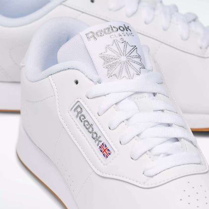 Close-up of Reebok Women's Princess Shoes, featuring a classic white design with the Reebok logo, signature Union Jack flag, star detail on the tongue, visible cushioned accents, and coordinating white laces and stitching.