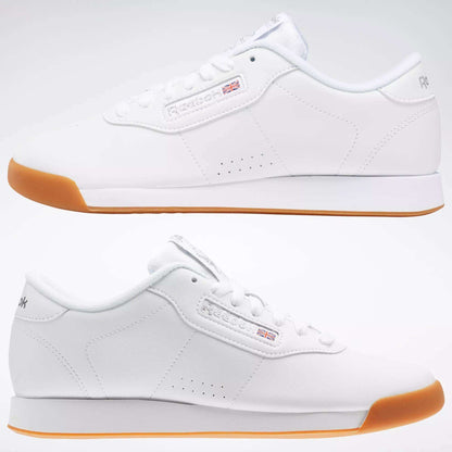 Two Reebok Women's Princess Shoes in white with gum soles sit side by side, facing opposite ways on a light background. Featuring simple stitching, soft cushioning, and the Reebok logo with a small British flag on the side.