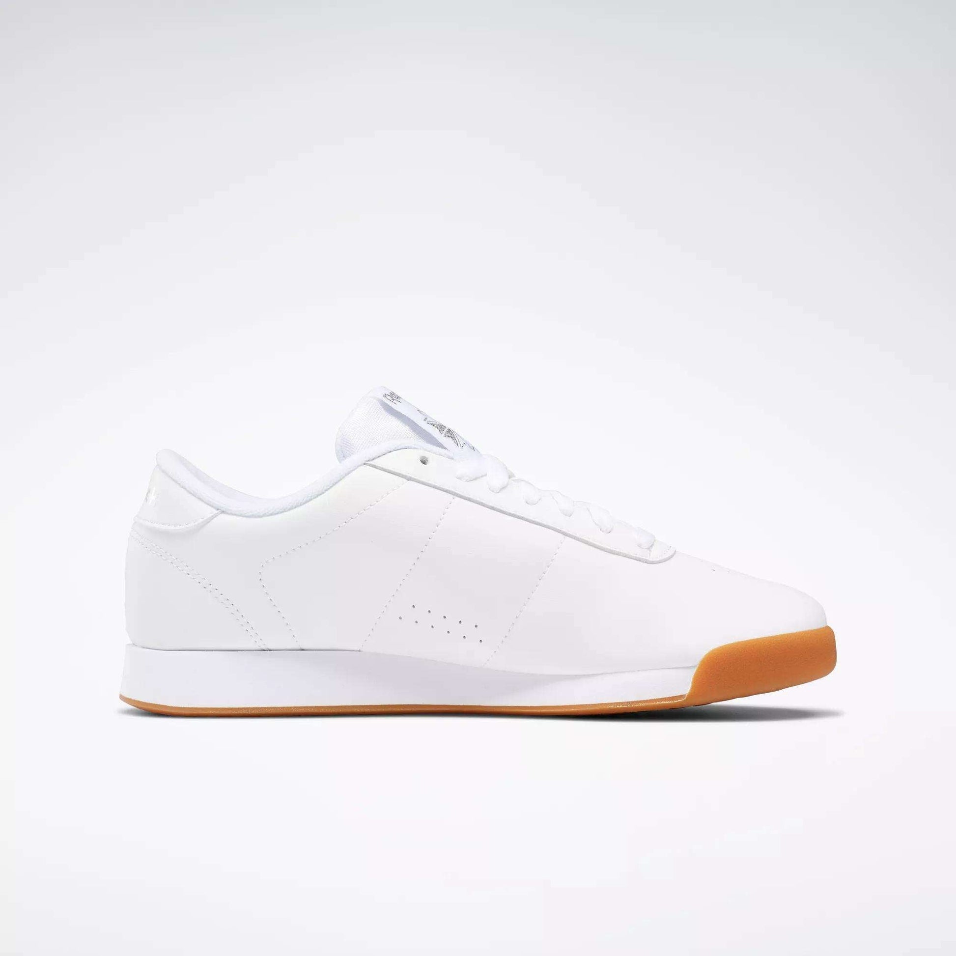 The Women's Princess Shoes by Reebok are classic white low-top sneakers with a minimal design, white laces, subtle stitching, and a cushioned light brown rubber sole, shown in profile against a plain light background.