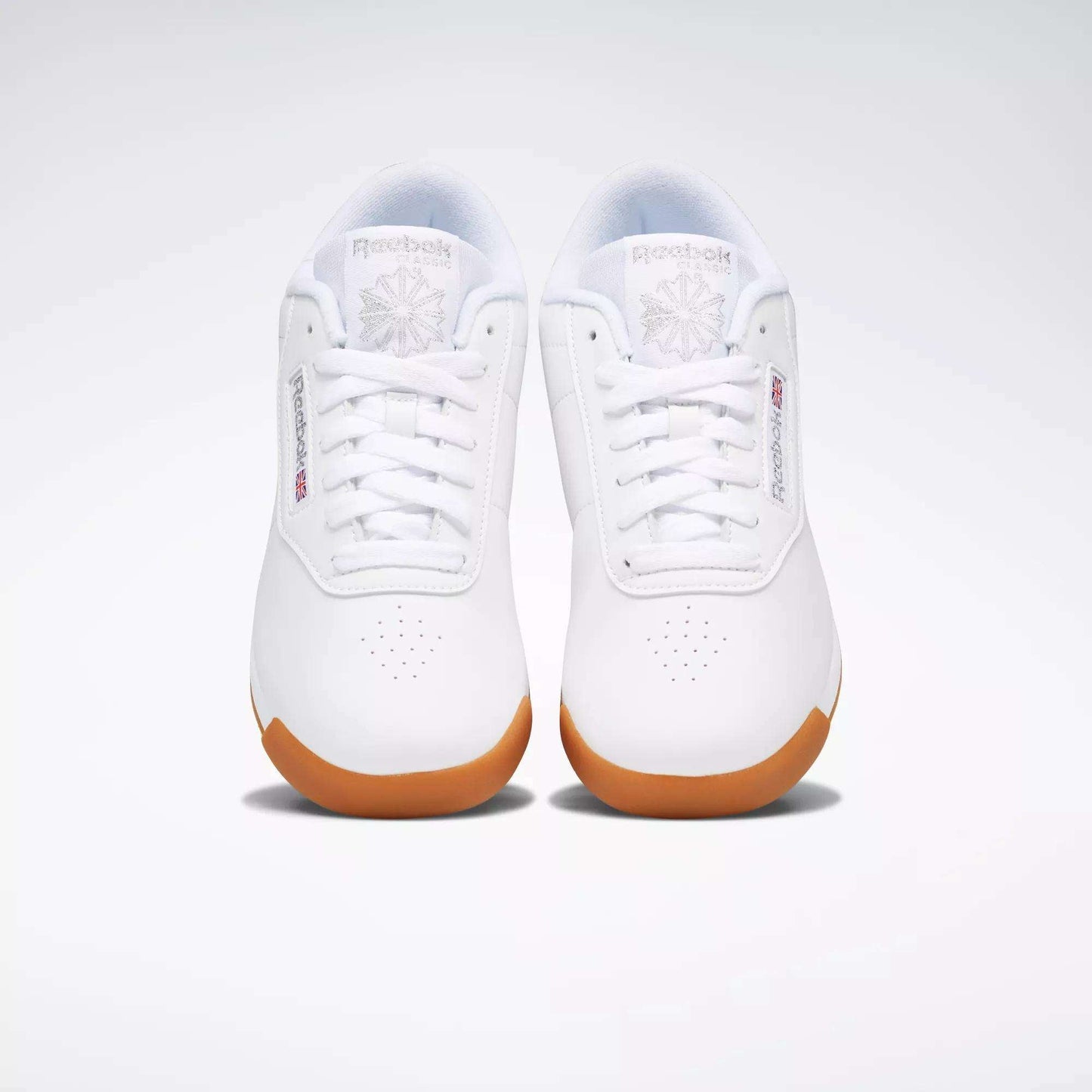A pair of Reebok Women's Princess Shoes in white with gum soles, shown from above on a plain light background. These sneakers feature Reebok logos, toe perforations, and provide excellent cushioning for all-day comfort.