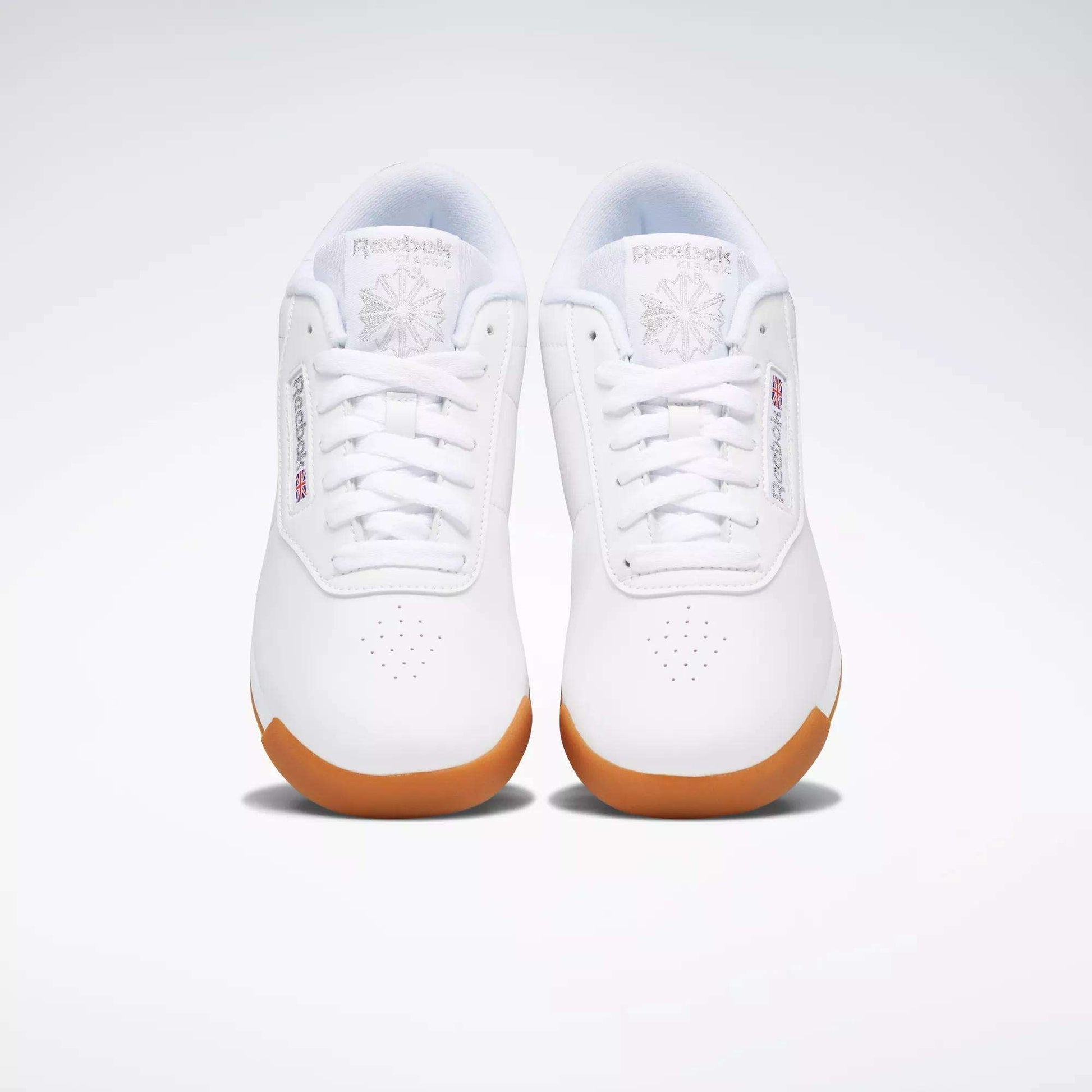 A pair of Reebok Women's Princess Shoes in white with gum soles, shown from above on a plain light background. These sneakers feature Reebok logos, toe perforations, and provide excellent cushioning for all-day comfort.