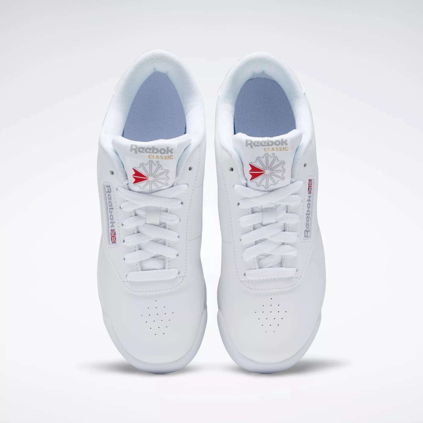 A pair of white Reebok Women's Princess Shoes viewed from above, featuring perforated toes, white laces, and classic Reebok branding with a red and gray logo—a timeless heritage style women’s sneaker.