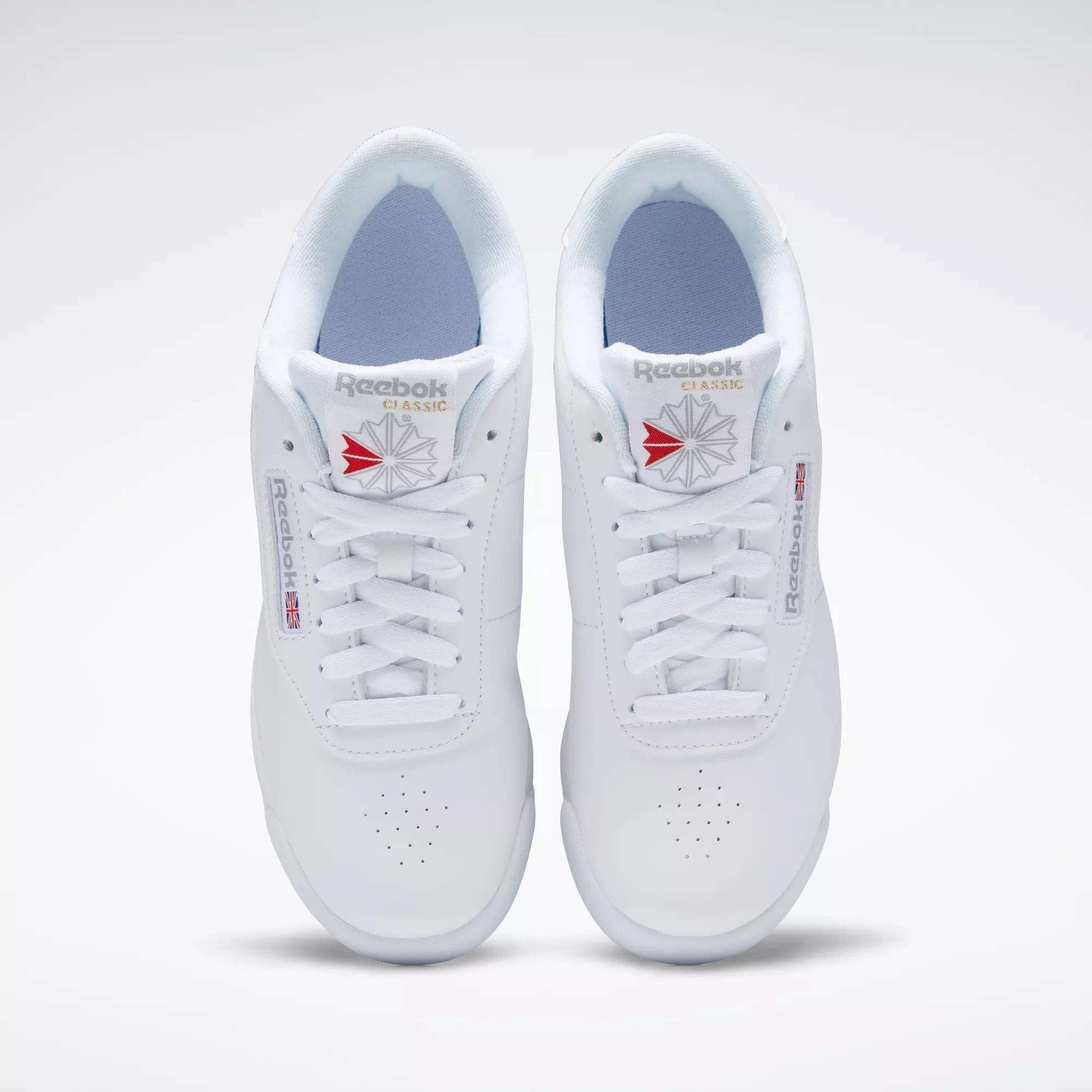A pair of white Reebok Women's Princess Shoes viewed from above, featuring perforated toes, white laces, and classic Reebok branding with a red and gray logo—a timeless heritage style women’s sneaker.