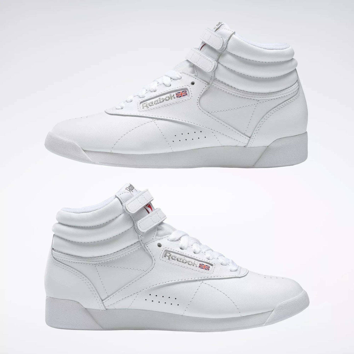 Two Reebok Women's Freestyle Hi Shoes in white are shown side by side on a light background, highlighting both outer and inner sides. Features include visible laces, velcro straps, and classic Reebok logos—ideal for women's fitness.