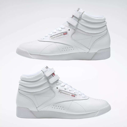 Two Reebok Women's Freestyle Hi Shoes in white are shown side by side on a light background, highlighting both outer and inner sides. Features include visible laces, velcro straps, and classic Reebok logos—ideal for women's fitness.