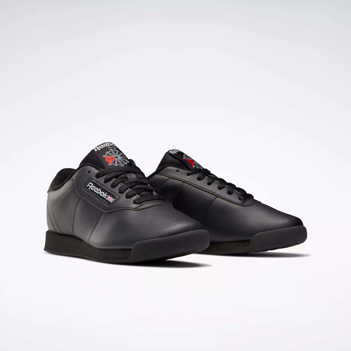 A pair of black Reebok Women's Princess Wide Shoes with black soles and laces, featuring the classic design and the Union Jack logo on the side and tongue, displayed on a plain light background.
