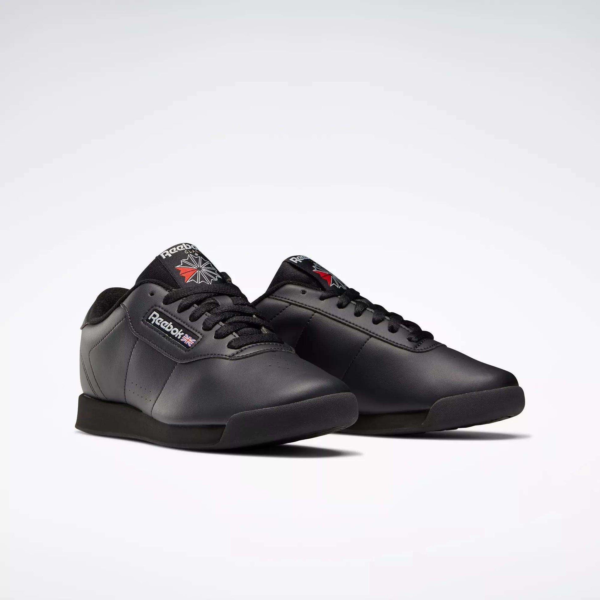 A pair of black Reebok Women's Princess Wide Shoes with black soles and laces, featuring the classic design and the Union Jack logo on the side and tongue, displayed on a plain light background.