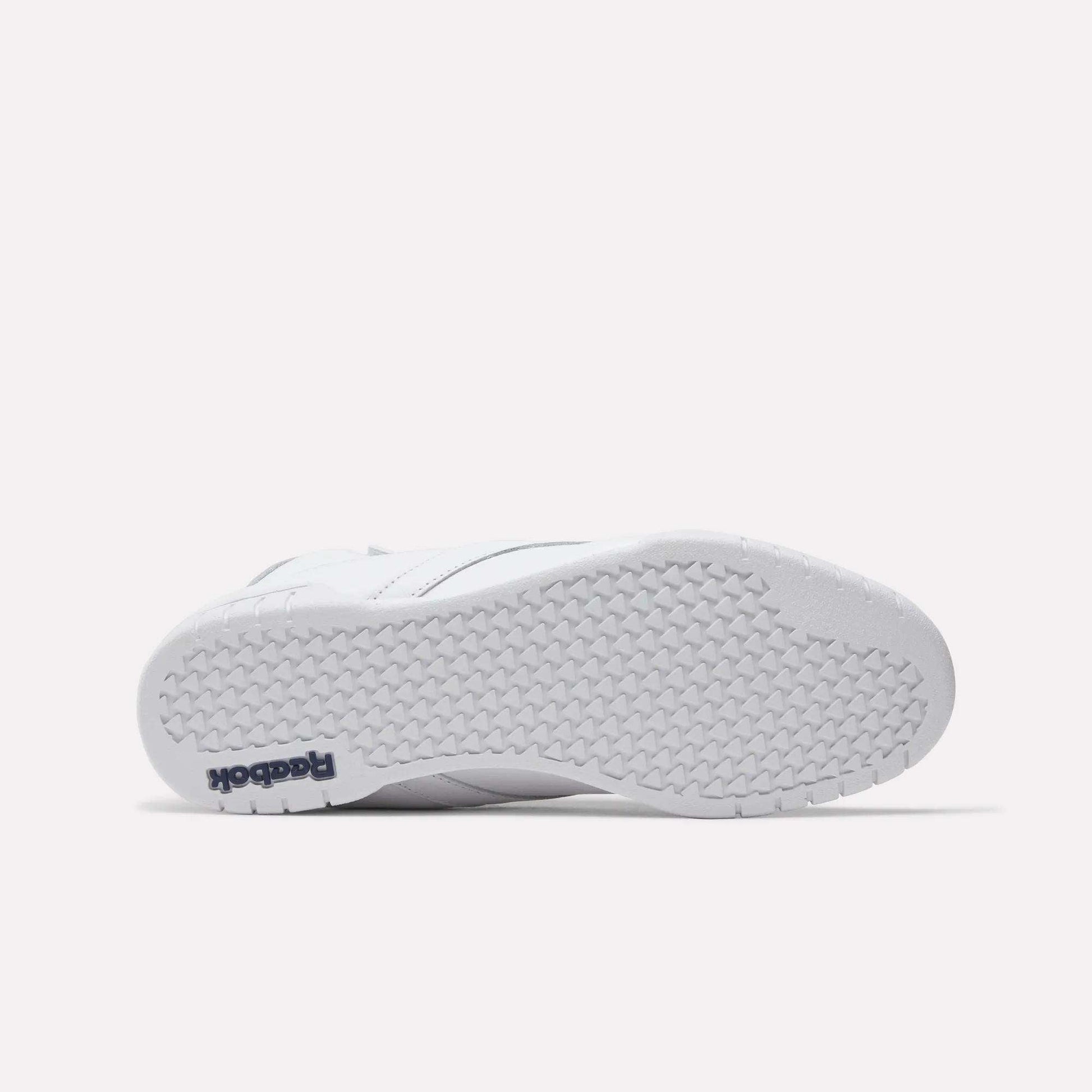 The image shows the bottom view of a white Reebok Men's EX-O-FIT Hi Shoe with a textured rubber sole. "Reebok" in navy blue appears on the heel, highlighting its classic old-school style.
