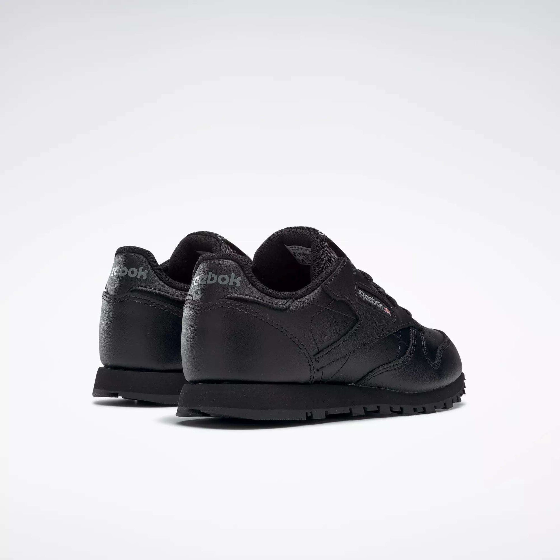 A pair of black Reebok Kids' Classic Leather Shoes - Preschool, shown from the back and side on a white background. These sneakers have a low-profile leather upper with Reebok branding on the heel and side.