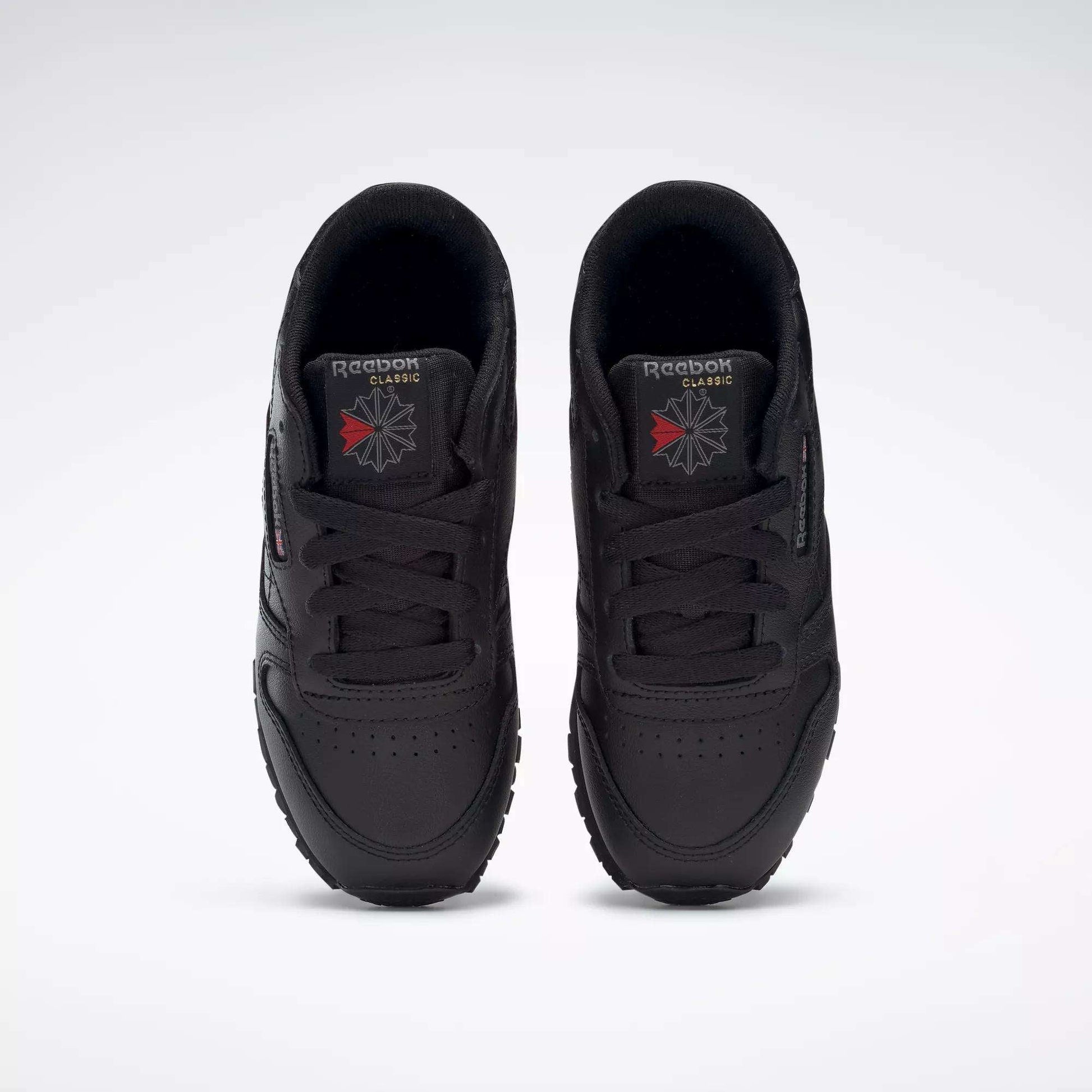 A pair of Kids' Classic Leather Shoes - Preschool by Reebok in black are shown from above, highlighting the leather uppers, laces, and Reebok logos on the tongues against a plain white background.