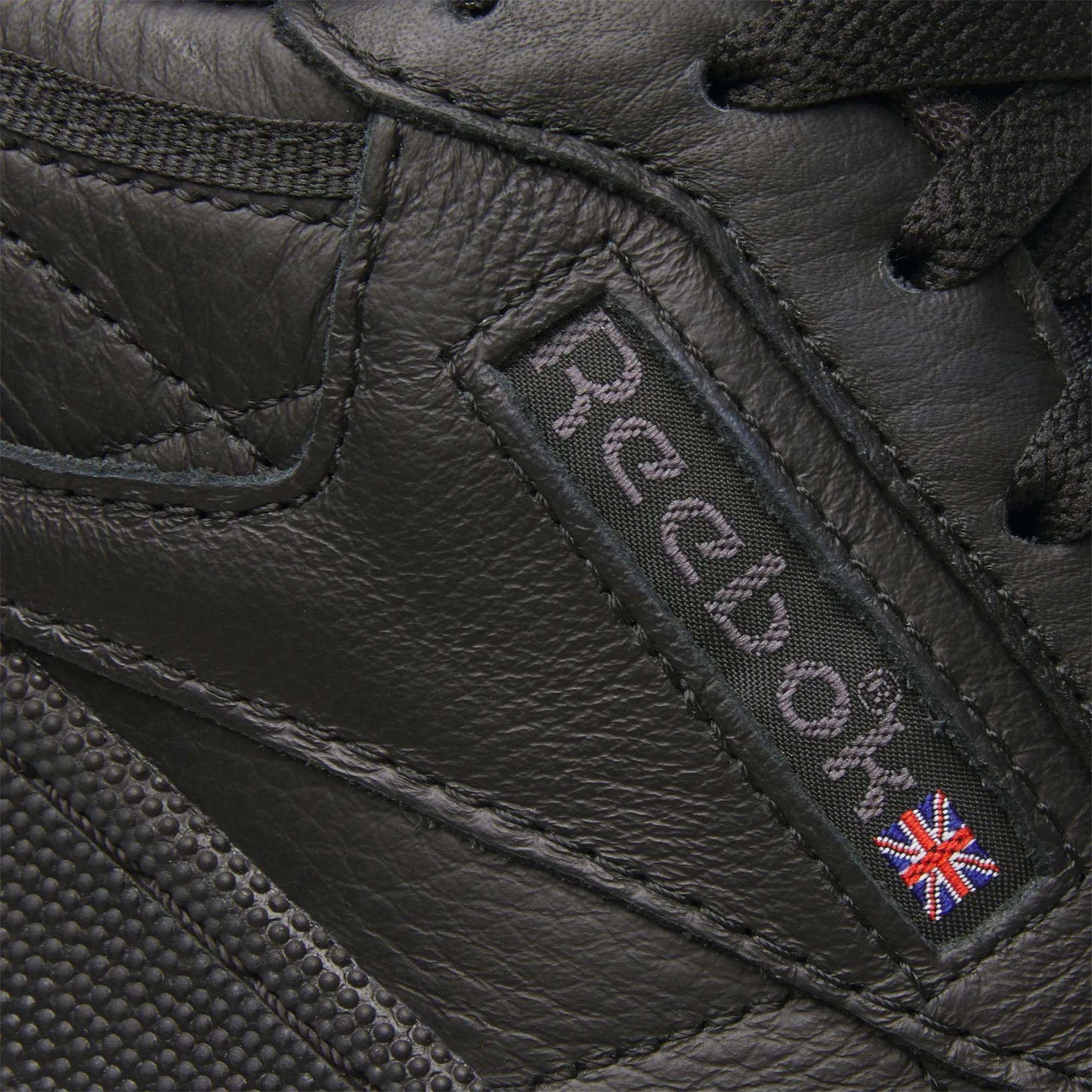 Close-up of the Club C 85 Shoes by Reebok, featuring black leather, the signature Reebok logo, and a small Union Jack flag patch stitched on the side—iconic details of this classic sneaker.