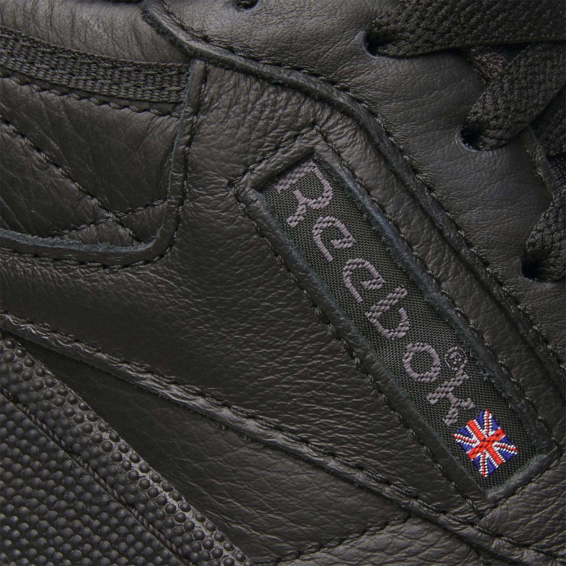 Close-up of the Club C 85 Shoes by Reebok, featuring black leather, the signature Reebok logo, and a small Union Jack flag patch stitched on the side—iconic details of this classic sneaker.