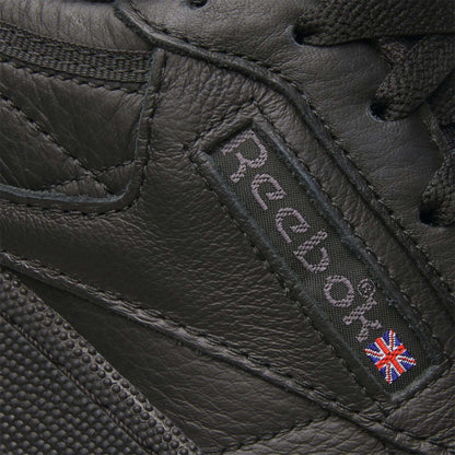 Close-up of the Club C 85 Shoes by Reebok, featuring black leather, the signature Reebok logo, and a small Union Jack flag patch stitched on the side—iconic details of this classic sneaker.