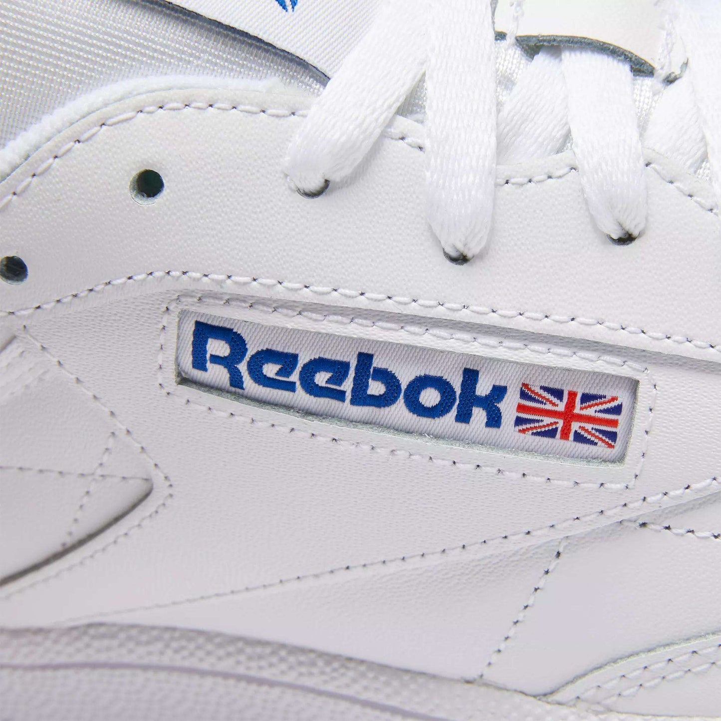 Close-up of Club C 85 Shoes by Reebok, featuring white leather, white laces, and a stitched blue Reebok logo with the British flag—classic tennis-inspired sneakers.