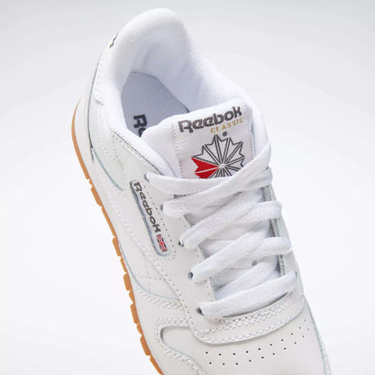 A close-up of Kids' Classic Leather Shoes - Preschool by Reebok, featuring a white leather upper, white laces, gum sole, Union Jack Reebok logo on the side, and a branded tongue with starburst design.