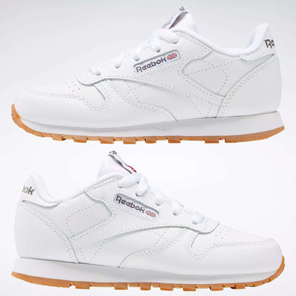 Two Kids' Classic Leather Shoes by Reebok in white are displayed side by side, showing the outer and inner sides. They feature a leather upper, Reebok branding, subtle stitching, and lightweight cushioning for comfort.